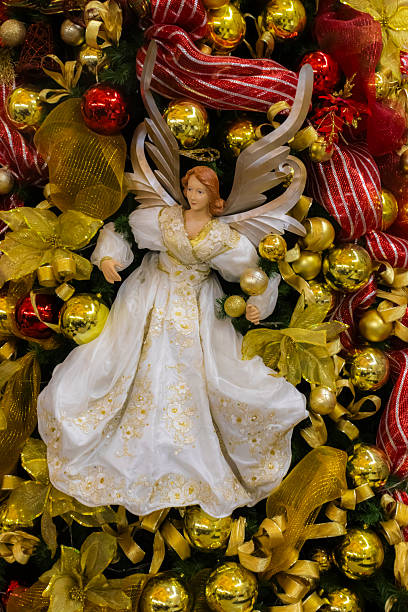 A beautiful Christmas angel ornament in a white and gold dress adorns a festive tree, surrounded by golden baubles and red ribbons, symbolizing peace and holiday spirit.