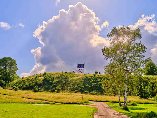 A serene Denmark landscape atop a gentle hill features a small white chapel with solar panels, a winding dirt path, and birch trees beneath a bright blue sky. The scene conveys calm, rural charm and the evolving light of late summer to autumn.