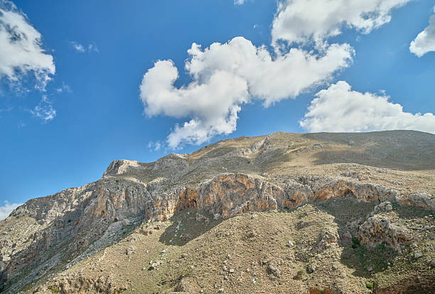 Crete landscape in Greece features a rugged mountain ridge, exposed rock, and dry, scrubby terrain beneath a vast blue sky dotted with fluffy clouds. This Greek scenery conveys rugged beauty, natural geology, and outdoor adventure, ideal for nature, travel, and landscape projects in Crete, Greece.