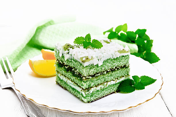 Piece of cake with mint and orange, white cream, confit, green sponge cake, sprinkled with white chocolate and green jelly in a plate, napkin on white wooden board background