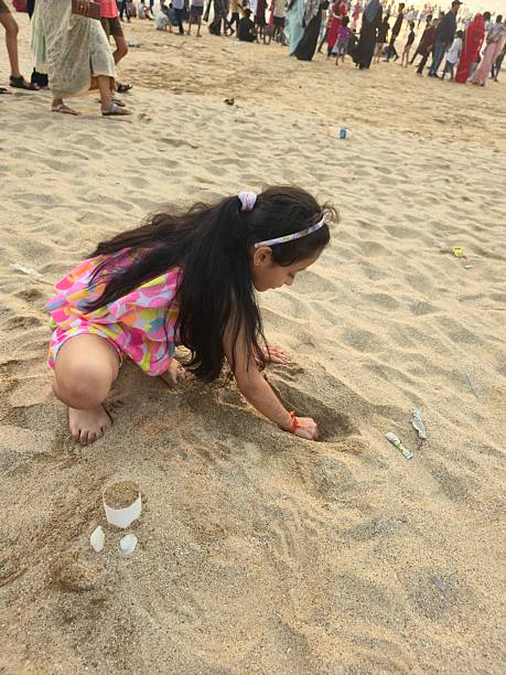 Young Indian girl sitting on sandy beach enjoying peaceful moment while playing with small cup filled with sand and seashell. Soft sunlight creating warm scene as child focuses on simple beach play with relaxed smile and gentle hand movements.