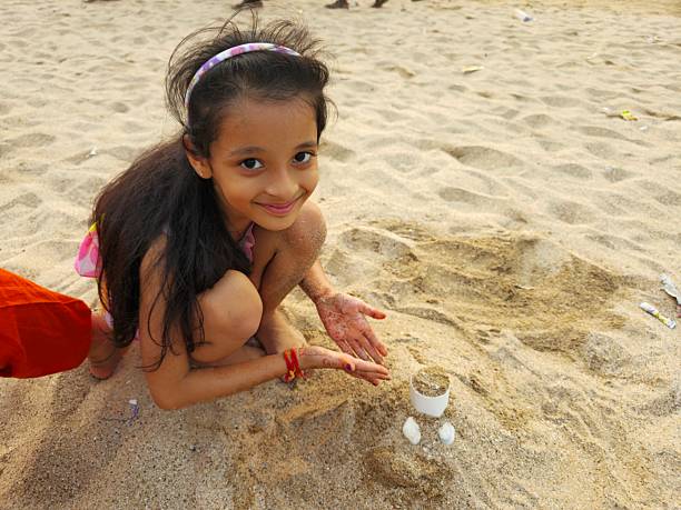 Young Indian girl sitting on sandy beach enjoying peaceful moment while playing with small cup filled with sand and seashell. Soft sunlight creating warm scene as child focuses on simple beach play with relaxed smile and gentle hand movements.