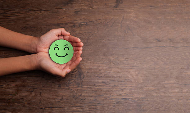 istock Hands gently holding a green happy face symbol on a wooden table background, representing positivity, emotional support, and mental health and wellness with space for text on the right. 2249863672