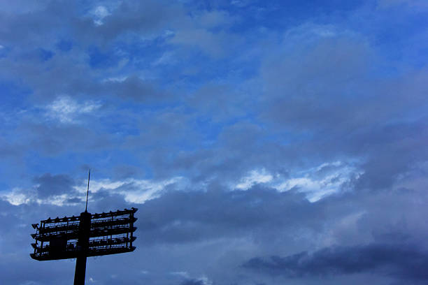 A stadium light stand is silhouetted against a dark, cloudy sky. The stand features multiple horizontal rows of lights and a vertical pole extending upwards. The background displays varying shades of blue, with dense cloud coverage creating a dramatic, dynamic sky.