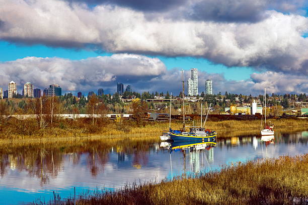 A sailing yacht anchored in the Fraser River Bay. The backdrop is the Vancouver shoreline, with its industrial zone and residential neighborhoods, and a cloudy sky.
