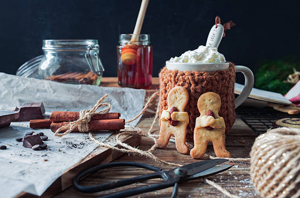 A festive winter scene features a front-facing mug of hot chocolate topped with whipped cream, nestled in a hand-knitted cozy. Two handmade almond cookies shaped like hugging figures lean against the mug, with rustic twine, cinnamon sticks, parchment paper, and chocolate chunks creating an authentic, sustainable Christmas mood. Copy space is available on the left and upper background.