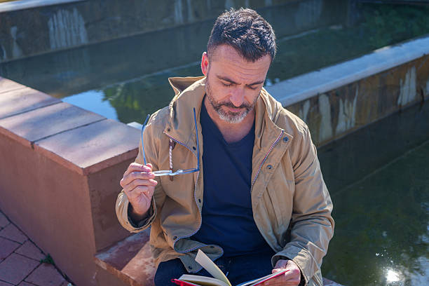 Thoughtful man reading a book, holding his glasses, and relaxing by a fountain. He is enjoying solitude and quiet reflection