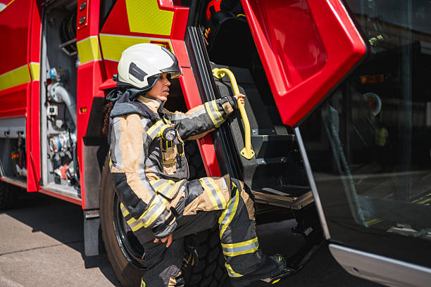 A young Hispanic female firefighter in protective gear steps out of a bright red fire truck at an airport, demonstrating readiness and professionalism in her critical role. The scene conveys action and dedication.