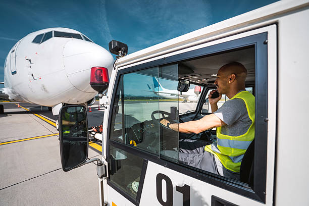 A Hispanic male in his 30s, seated inside a pushback tug, uses a radio device while operating airport ground equipment during daylight hours with maintenance and storage facilities in the background.