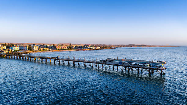 A tranquil coastal scene at Swakopmund, Namibia, featuring a long wooden pier extending over calm blue water with a town and distant dunes under a clear sunset sky.