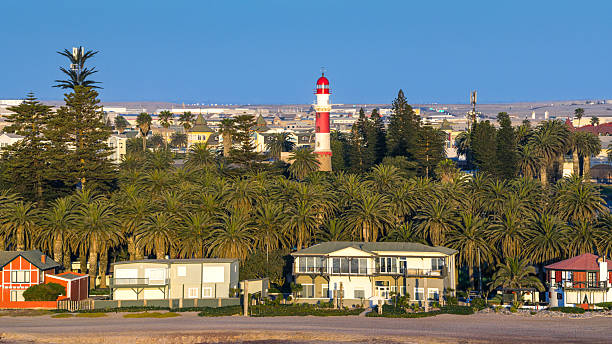 A coastal Swakopmund scene at golden hour featuring a red-and-white lighthouse behind dense palm trees, beachfront houses, and calm blue sky. Warm light creates tranquil coastal atmosphere.