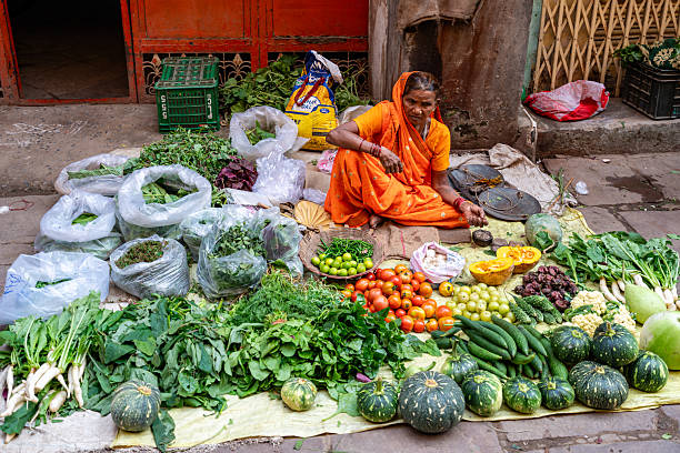 India. Uttar Pradesh state. Varanasi (Benares). A vegetable vendor sits behind her makeshift stall, selling fresh fruits and vegetables on a street in Varanasi (Benares). The woman wears a bright orange sari, a color often associated with spirituality and tradition in India