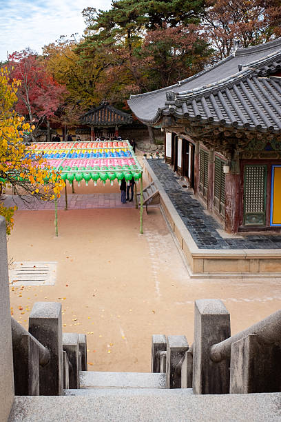 Gyeongju, South Korea November 13, 2025. Traditional Korean temple courtyard framed by fiery autumn foliage and ornate wooden architecture, paper lanterns hanging above stone paths as visitors stroll and pause in reverence