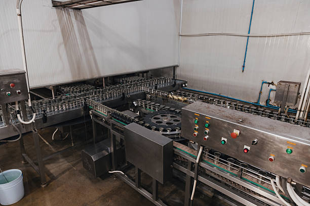 Workers operate a factory production line with machines moving items along a conveyor. Controls and equipment are set up around the workspace, reflecting a busy environment.