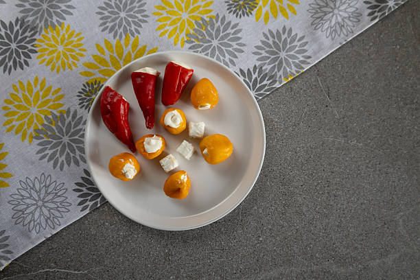 A plate of small stuffed red and yellow peppers is served alongside cheese cubes on yellow and gray patterned cloth and on gray background. The arrangement creates a simple, elegant appetizer presentation suitable for food, catering, or entertaining themes.