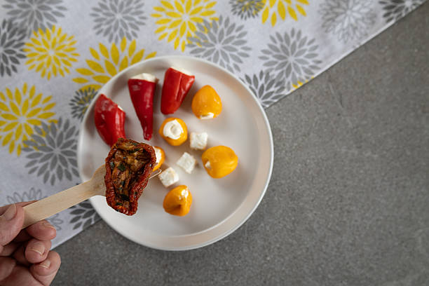A hand holding a wooden tasting stick with a stuffed dried tomato is positioned in front of a plate filled with red and yellow stuffed peppers and small cheese cubes. The colorful ingredients and textured surfaces create a fresh, homemade and inviting appetizer presentation. The combination of vegetables, cheese and rustic styling enhances the artisanal culinary mood.