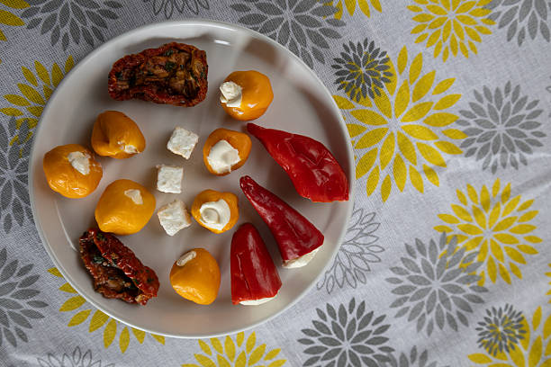 A plate of small stuffed red and yellow peppers and a roasted, seasoned tomatos is served alongside cheese cubes on yellow and gray patterned cloth. The arrangement creates a simple, elegant appetizer presentation suitable for food, catering, or entertaining themes.