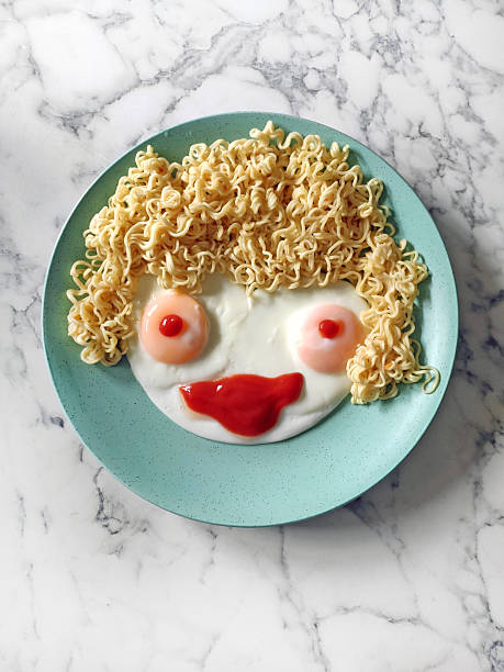 Creative food art on a turquoise plate: fried eggs with ketchup forming a face and instant noodles arranged as curly hair on a marble background.