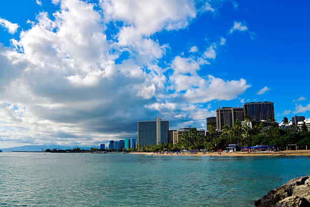 Honolulu, Hawaii, USA: the Waikiki Beach skyline stretching along the coast of Mamala Bay. The dense urban landscape features numerous high-rise luxury hotels and condominiums bordering the white sandy shoreline and the  waters of the Pacific Ocean. The architectural profile includes a mix of modern and mid-century designs, reflecting the area's development as a premier global resort destination.