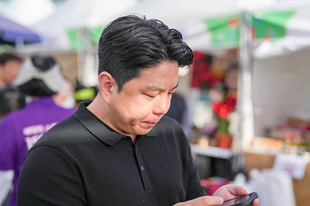 A man is looking at his cell phone while standing in a market. He is wearing a black shirt and he is focused on his phone