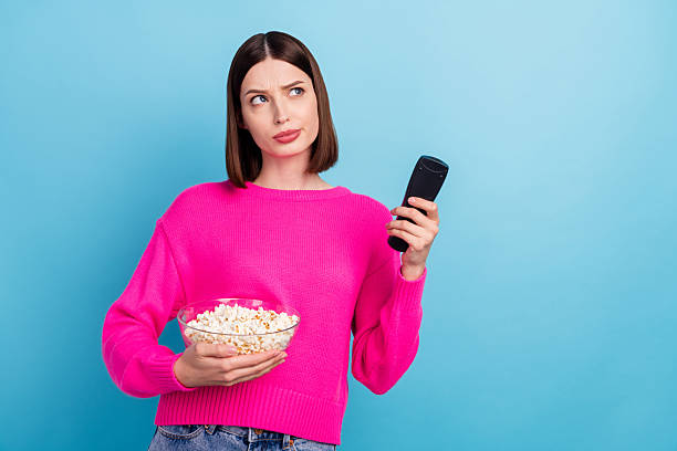 istock Photo of doubtful unsure young lady wear pink pullover choosing program looking empty space eating pop corn isolated blue color background 2249923745