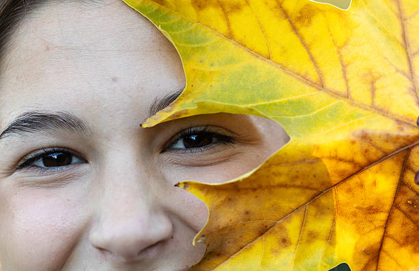 A girl smiles while holding a large yellow leaf close to her face. The background features natural greenery and bright autumn colors.