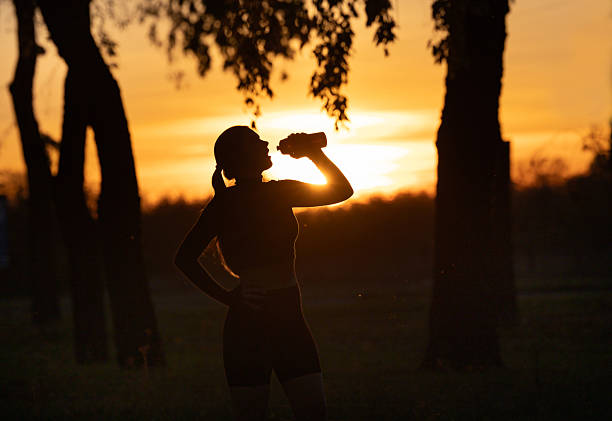 Runner drinks water during sunset, silhouetted by the warm orange sky and shadowy trees, capturing a peaceful moment after exercise