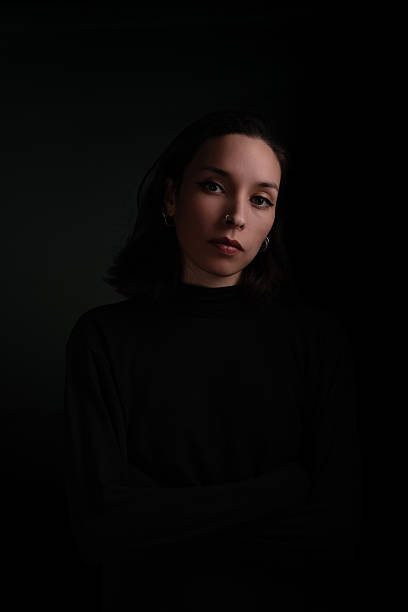A striking low-key portrait of a woman captured in a dark, minimalist studio setting. Soft and directional lighting highlights her facial features while the rest of the scene fades into shadow, creating an intimate and cinematic mood. Her calm, reflective expression enhances the emotional depth of the image, making it ideal for creative projects exploring themes of identity, introspection, elegance, or psychological narrative.