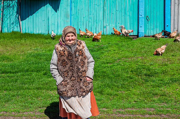 An older woman smiles while posing in front of her home with chickens grazing nearby