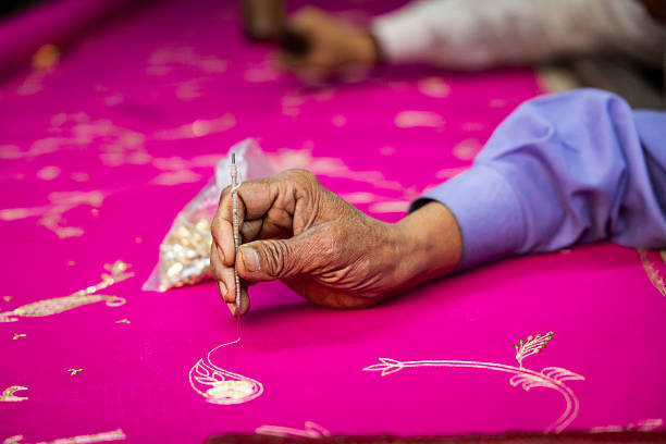 Close-up of a skilled hand holding a fine needle or awl to stitch delicate gold motifs onto bright magenta fabric. The image focuses on the weathered fingers and the intricate paisley and floral embroidery, with a small bag of beads or sequins blurred in the background.