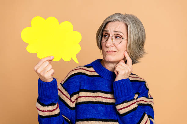 istock Mature woman with glasses holds a bright yellow speech bubble while thinking on a beige background 2249945004