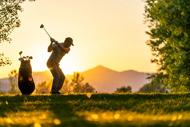 Mid-adult Caucasian male golfer captured in mid-swing with a driver, enjoying a competitive moment on the tee area of a sunlit golf course. The warm evening light enhances the scene, adding to the excitement of the game.