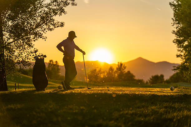 Mid-adult Caucasian male golfer standing on the tee box with a driver, enjoying a relaxed moment on the golf course as the sun sets behind the mountains, creating a serene atmosphere.