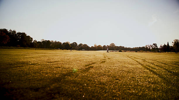 Caucasian mid adult couple enjoying a relaxed walk across a sunlit golf fairway, surrounded by lush greenery and open skies, during a pleasant afternoon. Their leisurely pace reflects a joyful and serene outdoor experience.