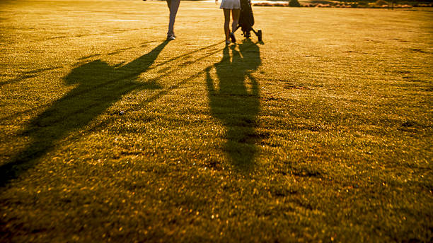 Caucasian mid adult couple enjoying a leisurely walk on a sunlit golf fairway. Their long shadows stretch across the well-maintained grass as they share a happy moment together, surrounded by the warm glow of the setting sun.