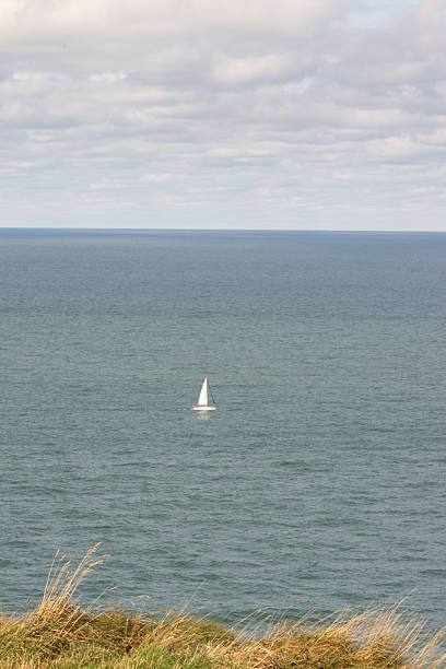 A solitary white sailboat drifting across the calm open sea below a cloudy sky, viewed from a grassy coastal cliff. Peaceful maritime landscape.