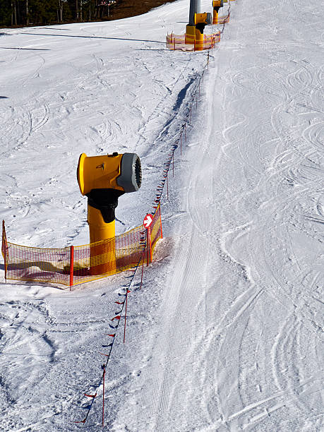 A high-angle photograph capturing a bright yellow snow cannon (snowmaking machine) positioned centrally on a wide, groomed ski slope. The machine is surrounded by orange safety netting and flags, marking the boundary of the piste. The background shows the freshly prepared white snow surface extending upwards, ensuring optimal skiing conditions. The image focuses on the technological infrastructure essential for modern winter sports resorts, highlighting themes of engineering, maintenance, and reliable snow production.