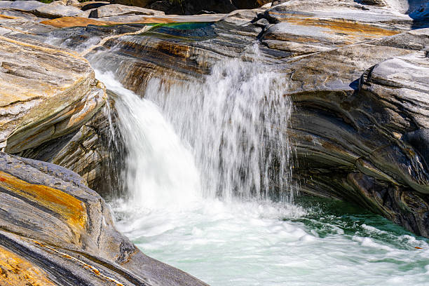 Vertical, close-up shot of white, churning river water cascading over colorful, layered metamorphic rock with streaks of orange and grey. The long exposure creates a silky effect on the rushing flow, highlighting the geological texture and raw power.