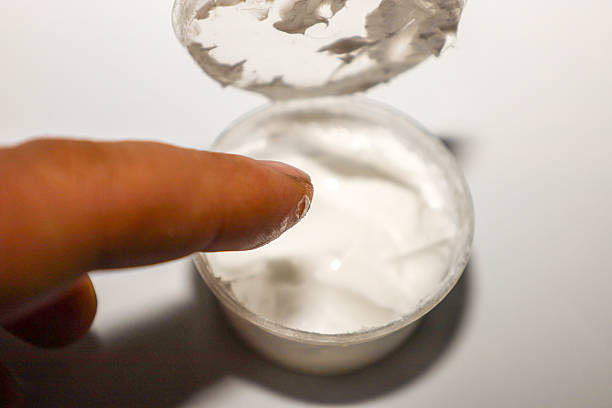istock ​A close-up shot shows a finger touching or dipping into a small, clear jar containing white cream or ointment, with the lid open above. 2249950598