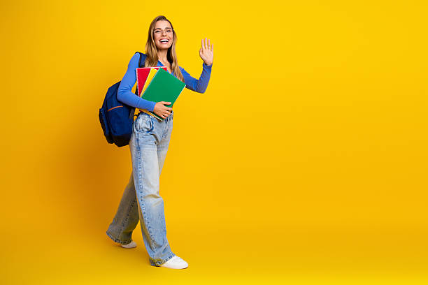 istock Young woman student with backpack waves hello while carrying colorful notebooks against a bright yellow background 2249970394