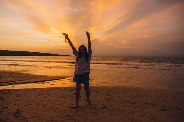 Happy mature female raising arms admiring bright dreamlike sunset walking near the reflective ocean in yellow and orange colours around at Jimbaran beach, Bali, Indonesia