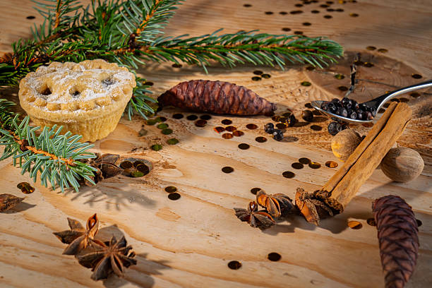 A golden-brown mincemeat pie featuring a decorative lattice pastry top is centered on a textured wooden table. Surrounding the pie are fresh green pine branches (Pinus sylvestris), reddish-brown pine cones, curved cinnamon sticks (Cinnamomum verum), star-shaped anise pods (Illicium verum), scattered juniper berries and black peppercorns (Piper nigrum), and gold confetti dots. The arrangement evokes a warm, festive holiday mood suitable for winter celebrations. This still life image can be used commercially in holiday recipe illustrations, Christmas baking articles, seasonal food packaging, festive table setting features, or winter-themed media content for authors and publishers.