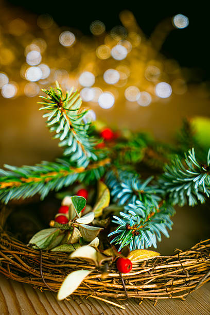 A close-up view of a handmade Christmas wreath featuring woven natural twigs as the base, adorned with fresh green evergreen branches from Picea species and clusters of bright red berries from Ilex aquifolium holly. The wreath rests on a textured wooden table surface, with soft golden bokeh lights blurred in the background to evoke holiday illumination. The composition highlights the textured needles, glossy leaves, and vibrant red accents against warm neutral tones. This image captures a cozy, traditional winter holiday mood suitable for commercial applications such as seasonal e-commerce product displays for wreaths and decorations, Christmas invitation designs, festive blog illustrations, editorial features on holiday crafts, and advertising for home goods or winter events. No people are present, focusing on the organic elements of the decoration in an indoor setting during winter.