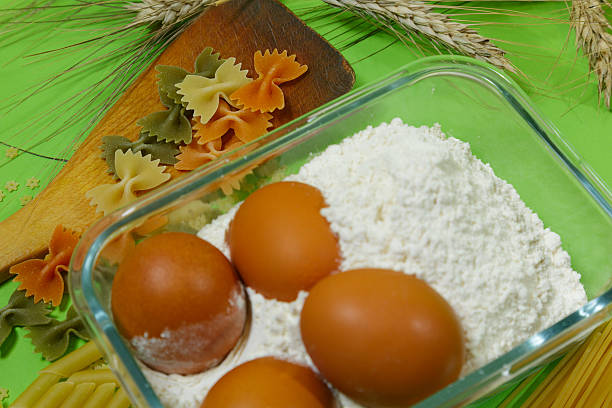 eggs resting in flour filled dish beside wooden spoon and farfalle, close tactile view of baking ingredients ready for dough