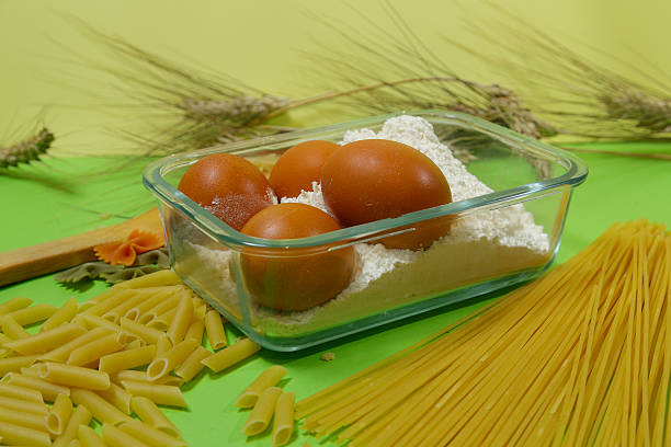 eggs nestled in flour filled dish among scattered penne and spaghetti, wheat stalks framing warm baking scene with tactile flour