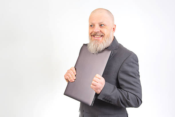 A cheerful bearded man in a grey suit holds a laptop, smiling confidently against a plain background, ideal for business and technology themes.