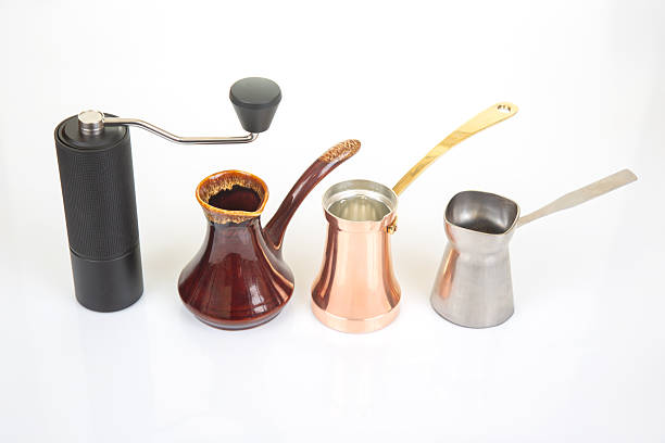 istock A black manual coffee grinder with a metallic handle stands beside a ceramic cezve, a copper cezve with a gold handle, and a stainless steel cezve on a white background, perfect for coffee brewing visuals 2249999457