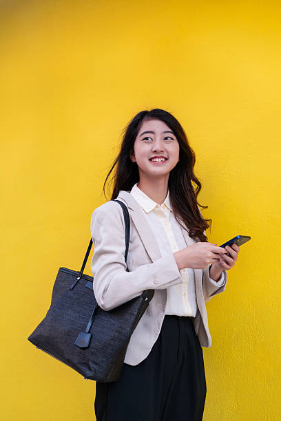 Young Asian woman using smart phone and standing against yellow colored background