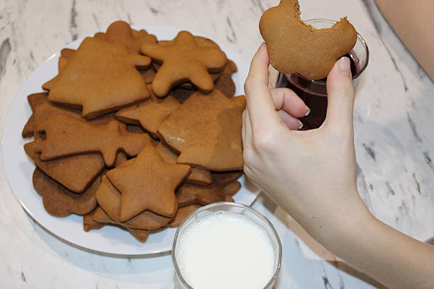 A hand holds a gingerbread cookie while sitting at a table. There is a plate stacked with more cookies and a glass of milk nearby.