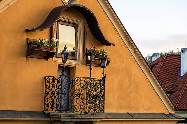 Vibrant yellow Prague facade featuring a decorative iron balcony, window box plants, and hanging lanterns. A charming urban detail capturing Czech architecture and warm, welcoming street life.